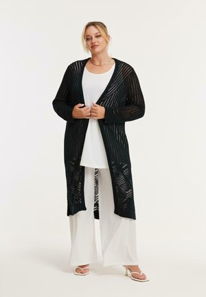 Kvinde iført lang sort åbenstrikket cardigan over hvid tunika og vidde benbukser, stående med hænderne foldet foran en lys baggrund.