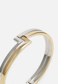 Pulsera bangle en tonos plateado y dorado con un diseño geométrico, que presenta una pequeña fila de cristales transparentes en el cierre para un detalle adicional.