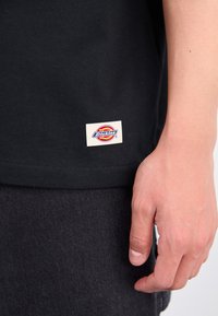 Rokas atpūšas pie gurniem, vilkusi melnas bikses un melnu kreklu ar mazu balto birku, uz kuras redzams zīmola Dickies logo.