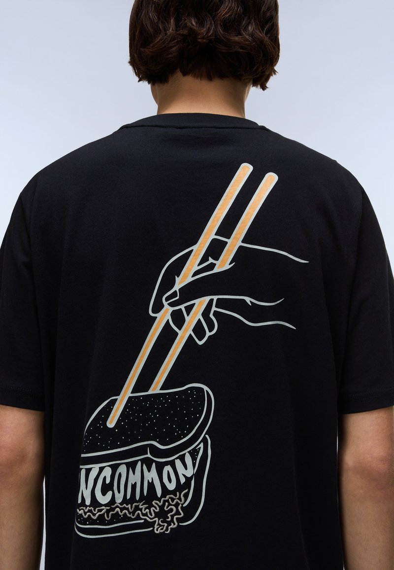 Camiseta de algodón negra con un gráfico en la parte posterior de una mano usando palillos sobre un rollo de sushi, con la palabra "POCO COMÚN" debajo.