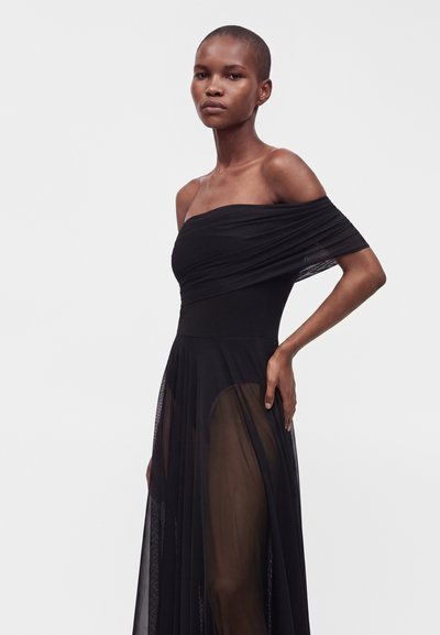 Femme aux cheveux courts portant une robe noire épaules dénudées avec un tissu transparent sur la jupe, debout devant un fond clair uni.