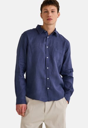 Chemise boutonnée à manches longues marine en tissu texturé avec une poche poitrine et des boutons blancs, portée avec un pantalon clair.
