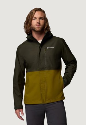 Mies, jolla on olkapäihin ulottuvat hiukset, pukeutuneena kaksiväriseen musta-oliivinvihreään Columbian takkiin, kädet osittain taskuissa.