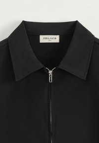 Chemise noire à col avec une fermeture éclair à l'avant. Fabriquée en tissu lisse. L'étiquette indique "FRILIVIN" avec la taille spécifiée. La fermeture éclair est dotée d'un matériel en métal.