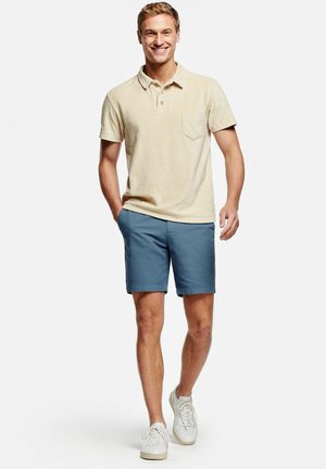 Lachende man loopt vooruit, draagt een beige kortemouwen poloshirt, blauwe korte broek en witte sneakers tegen een effen achtergrond.
