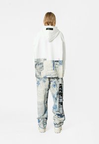 Sudadera blanca con patrones abstractos en azul y beige. Pantalones holgados con patrones similares y una etiqueta negra en el lado. Zapatillas blancas gruesas.