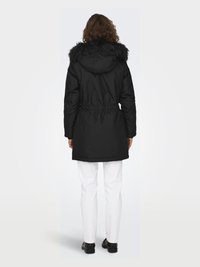 Black parka with a fur-lined hood, drawstring waist, and long sleeves. Worn over white pants, showing smooth texture and a relaxed fit.