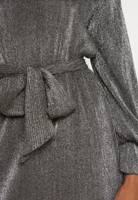 Skimrande, silvermetallisk klänning med en texturerad finish. Har en knytning i midjan och lång ärm, med vertikala ribbade linjer.