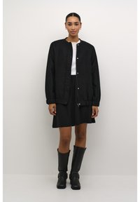 Čierna štruktúrovaná bomber bunda so cvokmi, nosená cez biely top a čiernu sukňu, doplnená čiernymi vysokými čižmami.