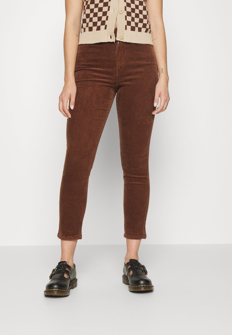 Personne portant un pantalon court en velours côtelé marron, des chaussures noires à boucle, et un haut sans manches à carreaux beige et marron avec boutons.