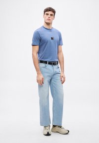 Modré bavlněné tričko s krátkými rukávy a čtvercovou černou nášivkou na hrudi, kombinované s světle modrými džíny s rovnými nohavicemi a béžovými teniskami.