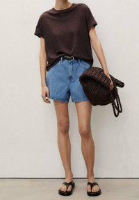 Personne portant un haut marron foncé à manches courtes, un short en jean bleu taille haute, des sandales noires, tenant un grand sac tressé foncé avec des poignées tressées.