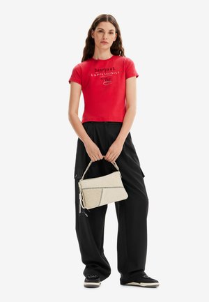 T-shirt rosso aderente con testo grafico, abbinato a pantaloni neri larghi. Una borsa a tracolla bianca tenuta in una mano, con un dettaglio con zip.