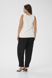 Weißes ärmelloses Top mit strukturierter Oberfläche, kombiniert mit lockeren schwarzen Hosen. Schwarze Sandalen mit Absatz und sichtbare Knöpfe an den Hosenbeinen.