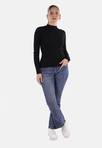 Top lungo nero a coste con collo alto, abbinato a jeans di denim blu e sneakers bianche. Il modello assume una posa neutra su uno sfondo chiaro.