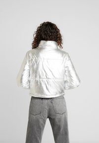 Sølvmetallic pufferjakke med høj krave og ribbet kant, der har en glat tekstur og quiltet design, båret med grå jeans.