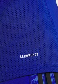 Close-up of breathable blue sports fabric with small ventilation holes and "AEROREADY" logo printed in white.