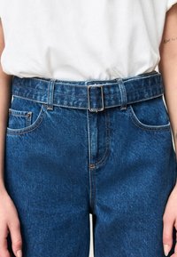 Denimbroek met een hoge taille, blauwe kleur, lussen voor een riem en een metalen gesp; voorzien van een klassieke vijfzakkenontwerp en contrasterende stiksels.