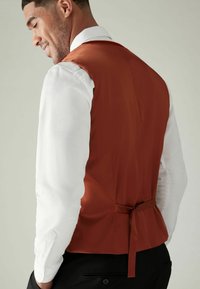 Gilet en satin marron au design ajusté, noué à la taille, porté par-dessus une chemise blanche texturée. Un pantalon noir est visible en dessous.