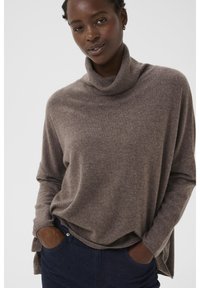 Suéter de cuello alto marrón hecho de un suave tejido de punto con un corte relajado y aberturas laterales, combinado con jeans de denim oscuro.