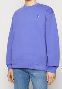 Sweatshirt in cotone viola con collo a giro, caratterizzato da un ricamo di un piccolo uccello blu sul lato sinistro del petto. Polsini e orlo a coste.