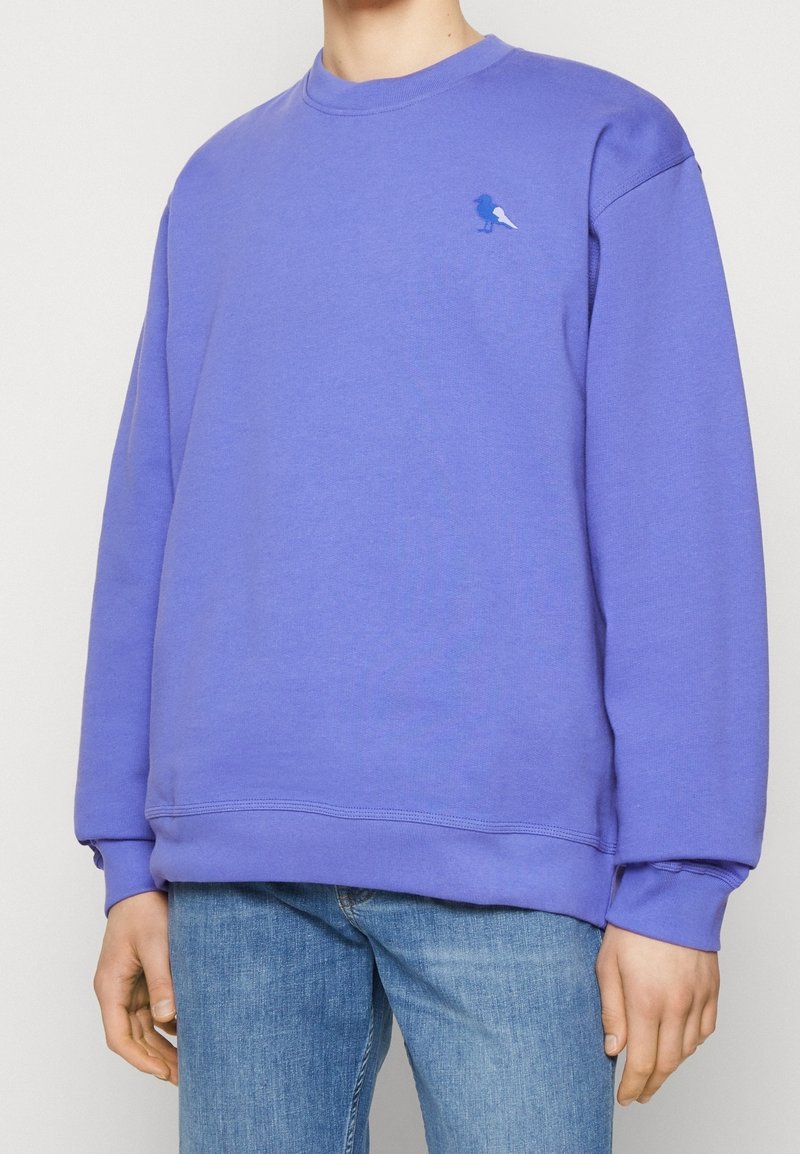 Sweatshirt in cotone viola con collo a giro, caratterizzato da un ricamo di un piccolo uccello blu sul lato sinistro del petto. Polsini e orlo a coste.