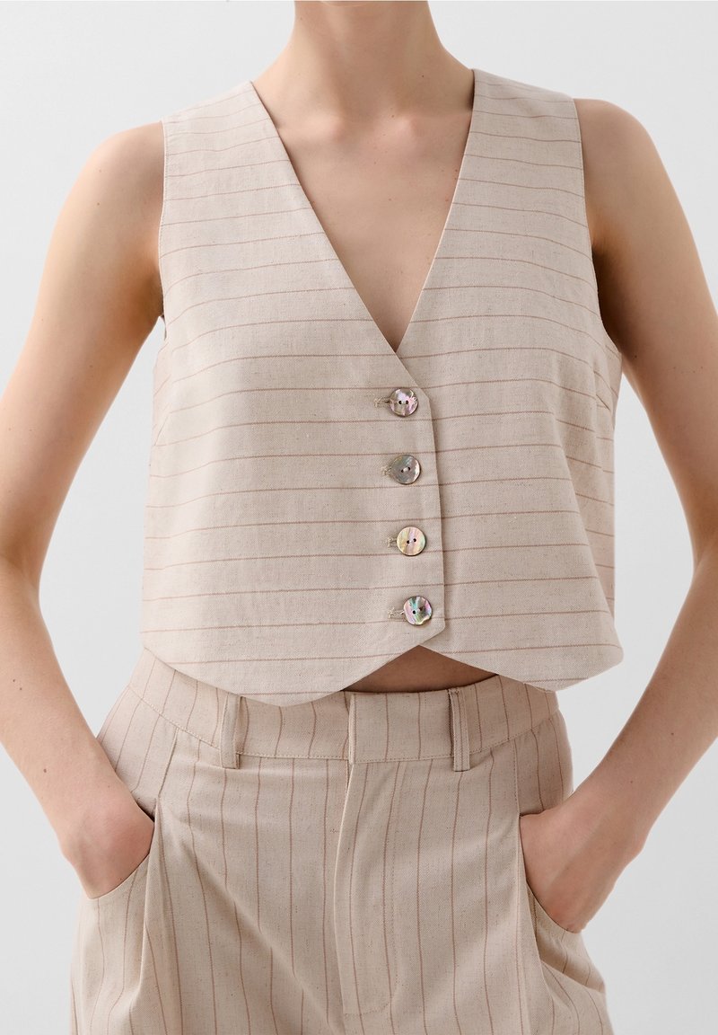 Veste beige sans manches à rayures fines boutonnée, associée à un pantalon taille haute assorti, les mains dans les poches, fond neutre.