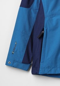 Chaqueta azul y marino con una tela texturizada, puños ajustables, bolsillo con cremallera y detalle del logo en la manga.