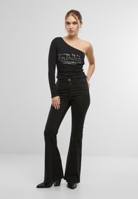 Svart en-armet topp med sølv "Von Dutch Originals" logo, kombinert med høyt belte svarte vide jeans og spisse svarte hæler.