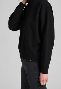 Persona con sudadera negra de manga larga y jeans gris oscuro, con la mano descansando cerca del cuello, de pie contra un fondo gris claro liso.