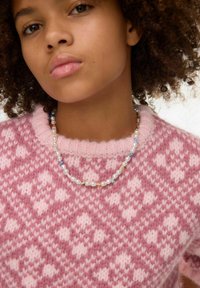 Rokaina rozā džemperis ar dimanta rakstiem, kura virsma ir mīksta un teksturēta. Delikāta pērlīšu kaklarota ar pērlēm un krāsainiem akcentiem.