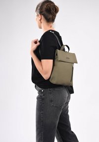 Mochila verde oliva con solapa plegable, bolsillo frontal con cremallera y correas ajustables negras, hecha de un material texturizado.