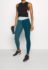 Czarny bezrękawnik z czerwonymi i niebieskimi akcentami, w połączeniu z turkusowymi legginsami z jaśniejszym pasem w talii. Czarne buty sportowe i butelka na wodę.