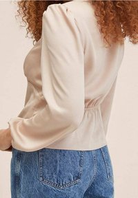 Femme aux cheveux bouclés portant un blouse en beige clair à manches longues et un jean en denim bleu taille haute, montrée de dos et de côté.