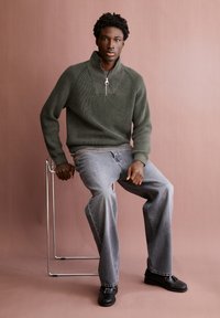 Junger Mann, der auf einem Metallhocker sitzt, trägt einen olivgrünen Halbzip-Pullover, graue Jeans und schwarze Quastenlober, vor einem schlichten braunen Hintergrund.