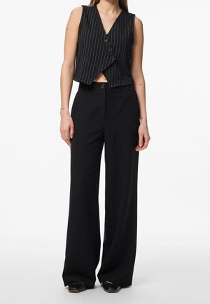Kobieta w czarnej kamizelce w prążki bez rękawów z guzikami oraz w czarnych, szerokich spodniach z wysokim stanem i czarnych butach, stojąca na białym tle.