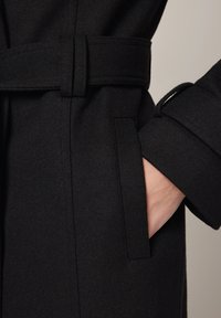 Czarny wełniany płaszcz z paskiem w talii, bocznymi kieszeniami i podwiniętymi mankietami. Tkanina ma gładką teksturę i elegancki krój.