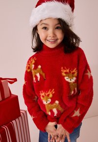Maglione rosso, morbido e peloso, con ricami di renne e stelle, colletto alto e texture soffice. Il modello indossa un cappello di Babbo Natale con pelliccia bianca.