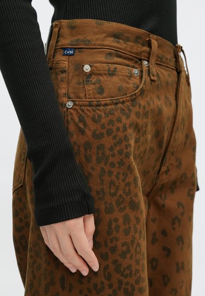 Jeans de ganga castanhos com padrão de leopardo preto, design de cinco bolsos e detalhes em metal prateado. Camisola preta de manga comprida com textura canelada visível.