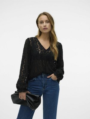 Blusa nera in pizzo con scollo a V, maniche lunghe e polsini a balze, abbinata a jeans in denim blu a vita alta e una pochette in pelle nera.