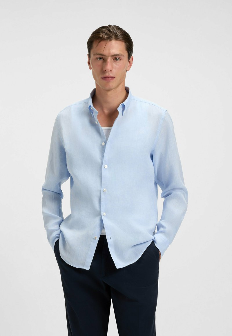 Jasnoniebieska koszula z guzikami uszyta z miękkiego, lekkiego materiału, z szerokim kołnierzem, długimi rękawami i kieszeniami z przodu, noszona na ciemnych spodniach.