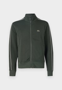 Chaqueta verde con cremallera fabricada con tela suave. Cuello alto, puños acanalados, bolsillos laterales y detalles blancos en las mangas. Logotipo de Lacoste incrustado.