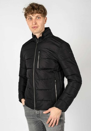 Chaqueta acolchada negra con un cuello alto, textura acolchada, cremallera frontal y dos bolsillos laterales, combinada con jeans gris claro.
