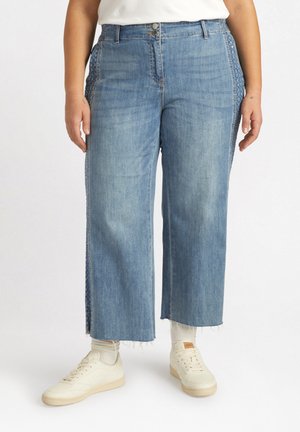 Weite blaue Jeanshose mit fransigem Saum, Knopfverschluss und dekorativem Seitenpanel-Design, kombiniert mit weißen Sneakers.