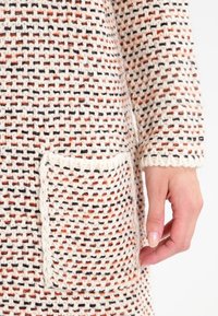 Trikåtyg i krämfärgad med svarta och orange sprickor; har en texturerad ficka; hand synlig som håller plagget.