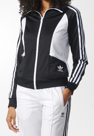Veste de survêtement Adidas noire et blanche avec fermeture éclair à l'avant et logo sur la poche, associée à un pantalon de survêtement blanc Adidas avec des bandes noires.