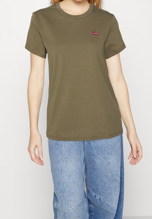 Person trägt ein olivgrünes Levi's T-Shirt und lockere hellblaue Jeans aus Denim vor einem schlichten weißen Hintergrund.