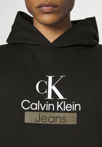 Närbild på en person som bär en svart hoodie med vit och guldfärgad "cK Calvin Klein Jeans"-logotyp tryckt på bröstet.