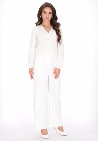 Fehér overál V-nyakkivágással, hosszú ujjak és elöl gombok. Széles szárú nadrágokkal és sima anyagból készült. Fehér magassarkúval viselve.