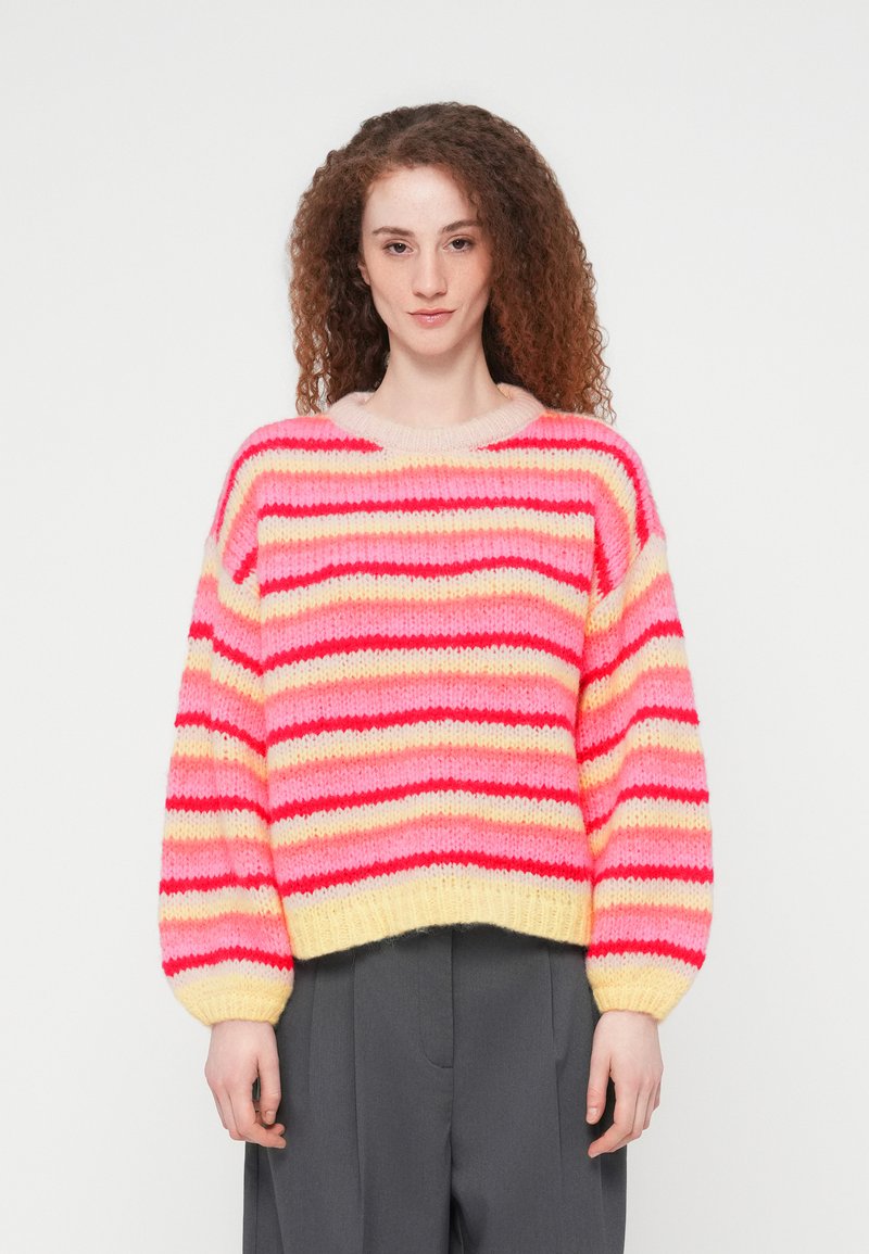 Γυναίκα με καστανά σγουρά μαλλιά, φορώντας ριγέ πλεκτό πουλόβερ σε ροζ, κόκκινο και κίτρινο χρώμα και σκούρο γκρι παντελόνι, στέκεται μπροστά σε λευκό φόντο.